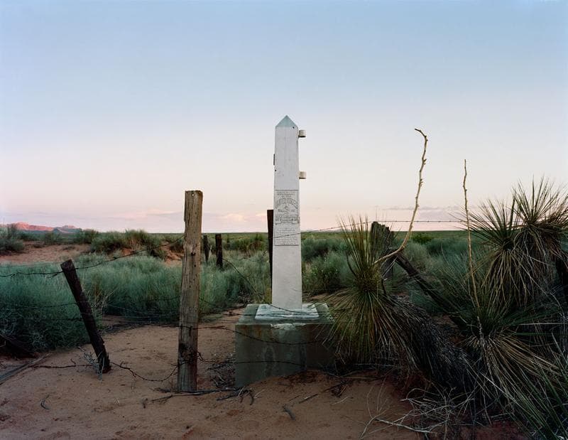 Photograph of an obelisk seen at dusk in a desert landscape, with a barbed-wire fence in the foreground