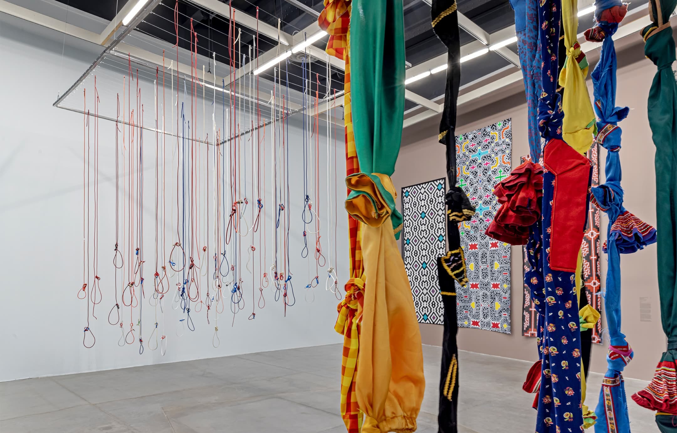 Photograph of a tall art gallery with two artworks suspended from the ceiling, one of which is a composition of multicolored textiles knotted together and hanging in long strands, the other of which appears to be colored ropes with looped shapes at the bottom. In the background, through the two foreground works, we can see three large hanging paintings with graphic motifs in black and white with spots of color.