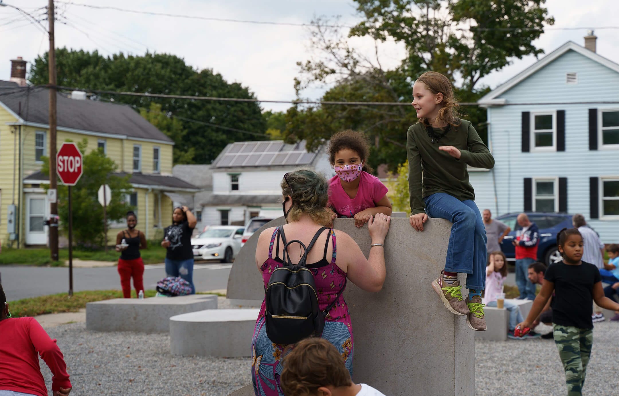 Several kids climbing on a concrete form in a busy playground; a woman stands near them with her back to the camera