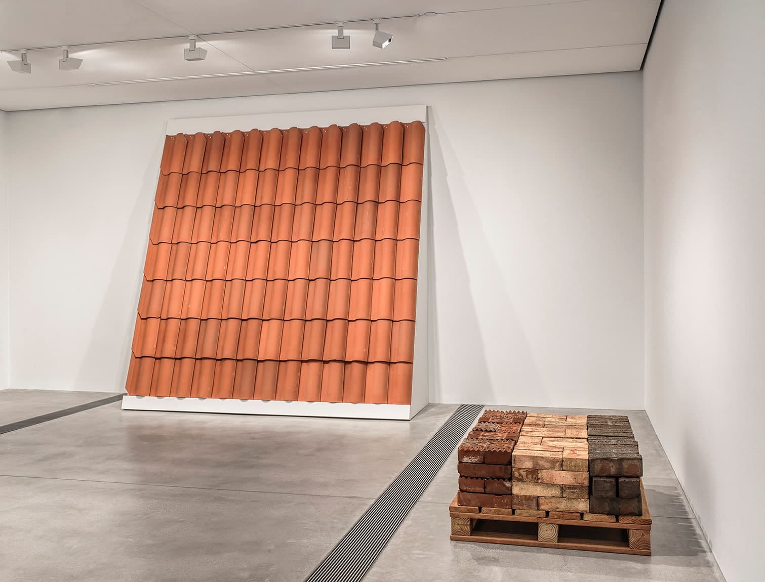 Photograph of a pallet of bricks and a tall section of terracotta roof tiles in a white-cube gallery