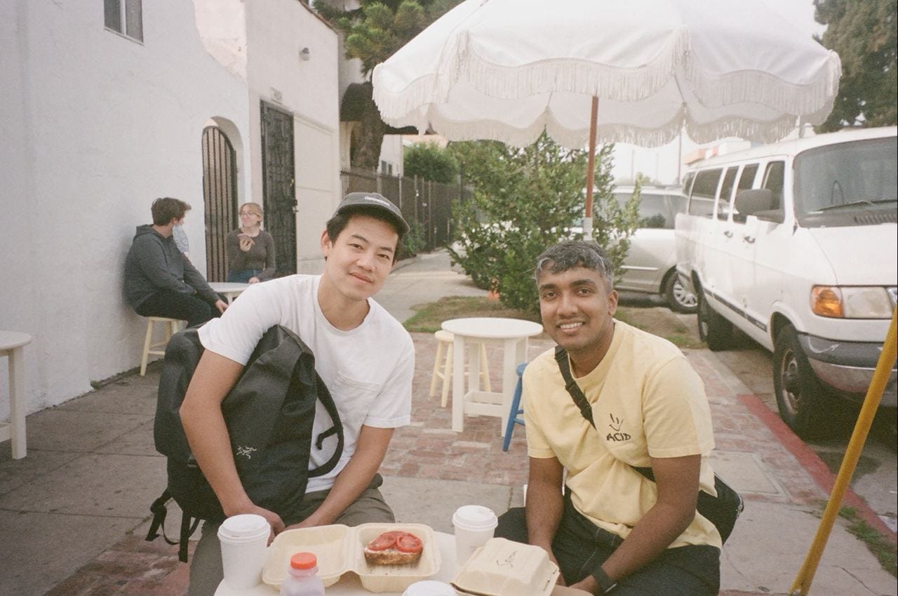 Portrait of Andy Chung and Mehdi Mulani sitting at an outdoor table with food and coffee cups; other tables, diners, and some nearby parked cars are in the background
