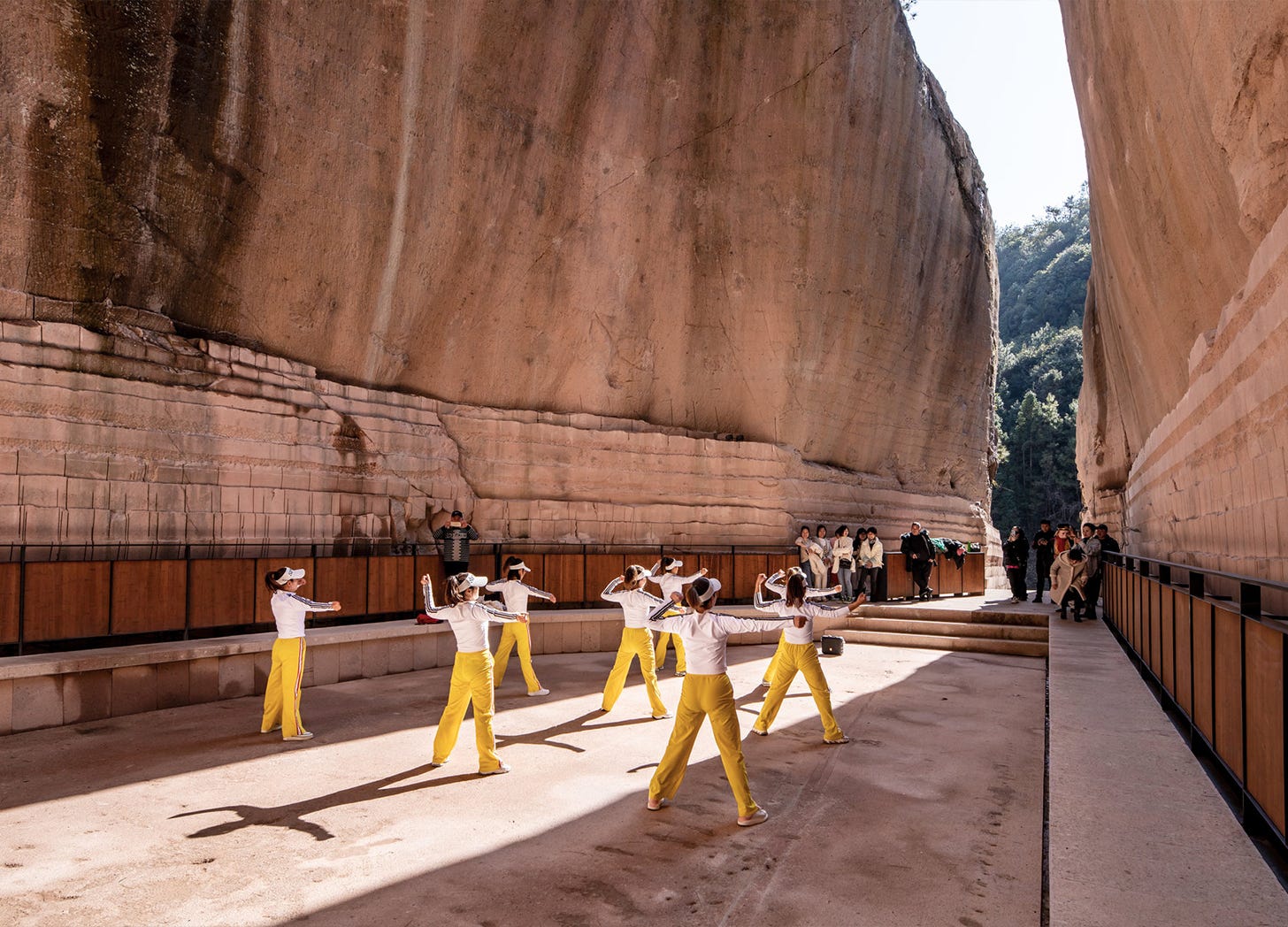 Dancers in yellow pants and white shirts perform on a recessed rectangular "stage" set within a rock quarry; beyond them, visitors watch the performance and a narrow opening reveals trees and a hill in the background